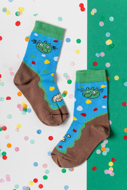 Blaue Olchis Kindersocken mit grünen Olchi-Gesichtern und brauner Sohle auf einem Hintergrund mit buntem Konfetti.