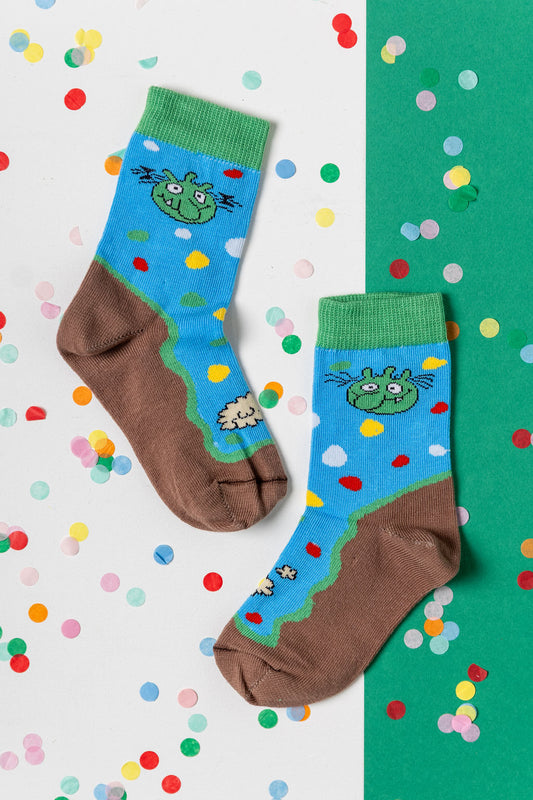 Blaue Olchis Kindersocken mit grünen Olchi-Gesichtern und brauner Sohle auf einem Hintergrund mit buntem Konfetti.