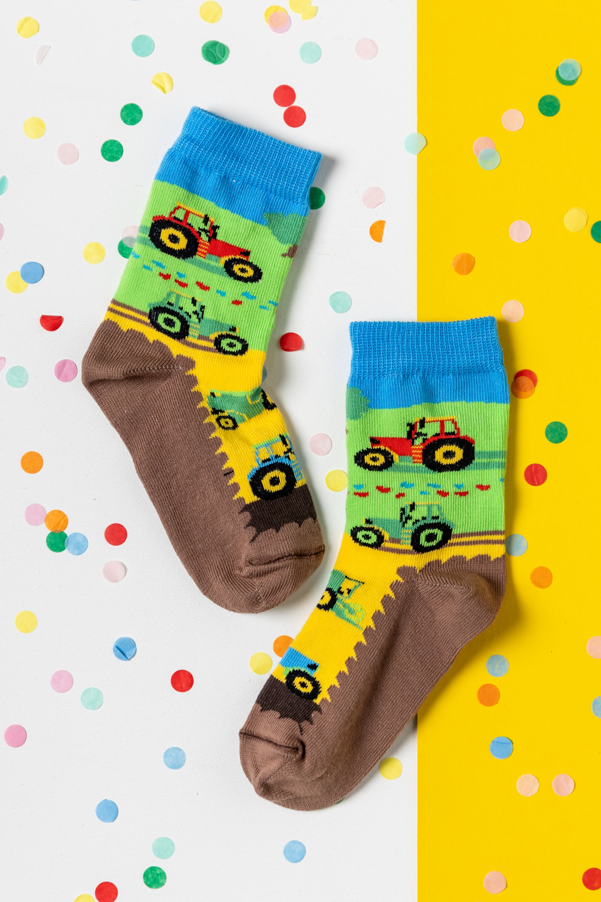 Bunte Kindersocken mit roten und grünen Traktoren auf einer Wiese mit blauem Himmel, dekoriert mit Konfetti.