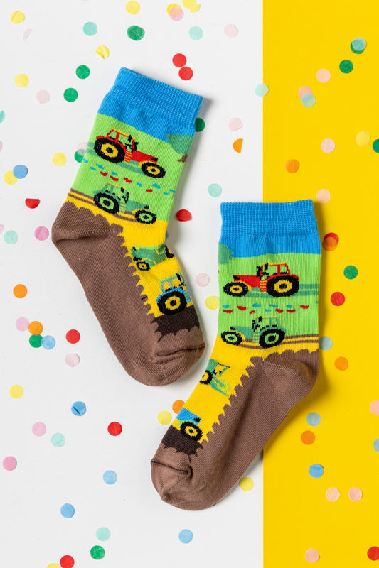 Bunte Kindersocken mit roten und grünen Traktoren auf einer Wiese mit blauem Himmel, dekoriert mit Konfetti.
