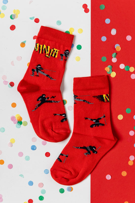 Zwei rote Kindersocken mit schwarzen Ninja-Motiven auf einem Hintergrund mit buntem Konfetti.